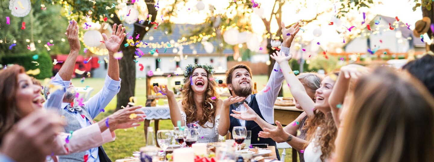 Eine Hochzeit im Garten, das Brautpaar sitzt am Kopfende des Tisches, links und rechts sitzen Gäste, Konfetti fliegen durch die Luft
