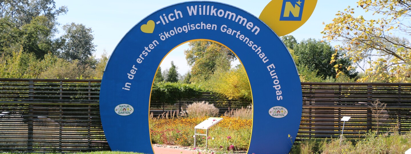 Ein blau-gelber Torbogen, der als Eingang zur GARTEN TULLN dient. Auf dem Bogen ist zu lesen: "Herzlich Willkommen in der ersten ökologischen Gartenschau Europas", hinter dem Torbogen blühen Blumen.