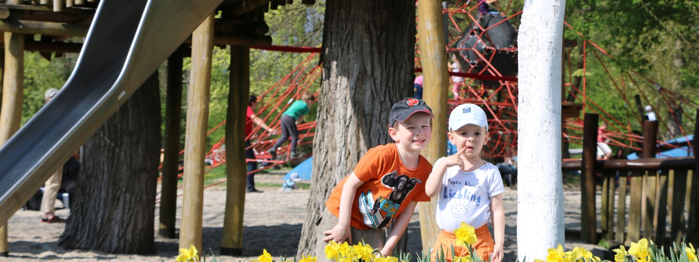 Zwei Kinder vor der Rutsche auf dem Abenteuer- und Naturspielplatz, vor ihnen blühen gelbe Narzissen