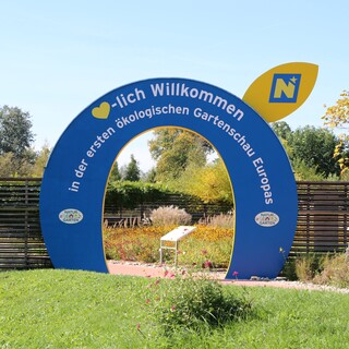 Ein blau-gelber Torbogen, der als Eingang zur GARTEN TULLN dient. Auf dem Bogen ist zu lesen: "Herzlich Willkommen in der ersten ökologischen Gartenschau Europas", hinter dem Torbogen blühen Blumen.