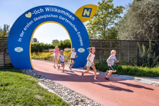 Fünf Kinder laufen durch einen Torbogen. Auf diesem ist zu lesen: "Herzlich Willkommen in der ersten ökologischen Gartenschau Europas"